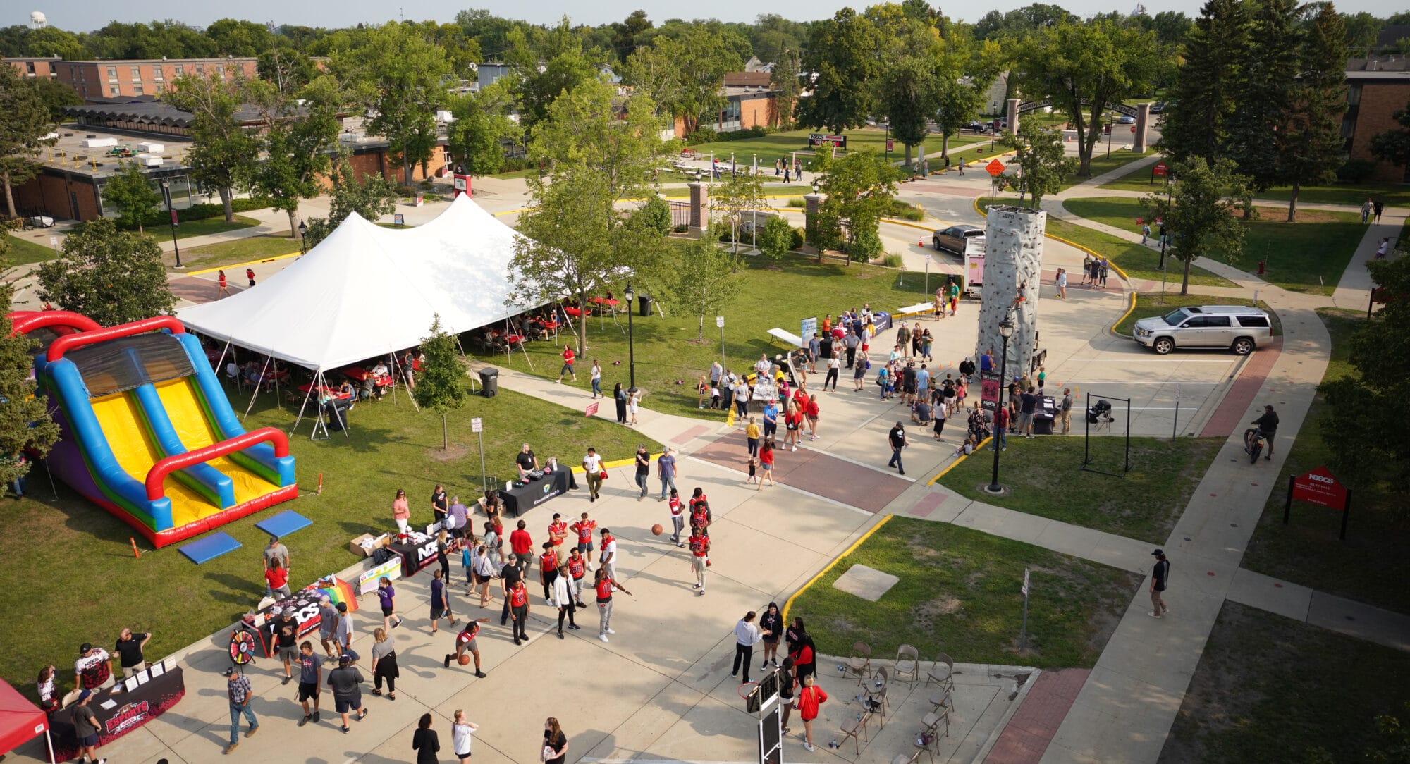 Aerial view of a lively outdoor campus event with a large white tent, inflatable slide, rock climbing wall, vendor tables, and crowds of students and staff participating in various activities.
