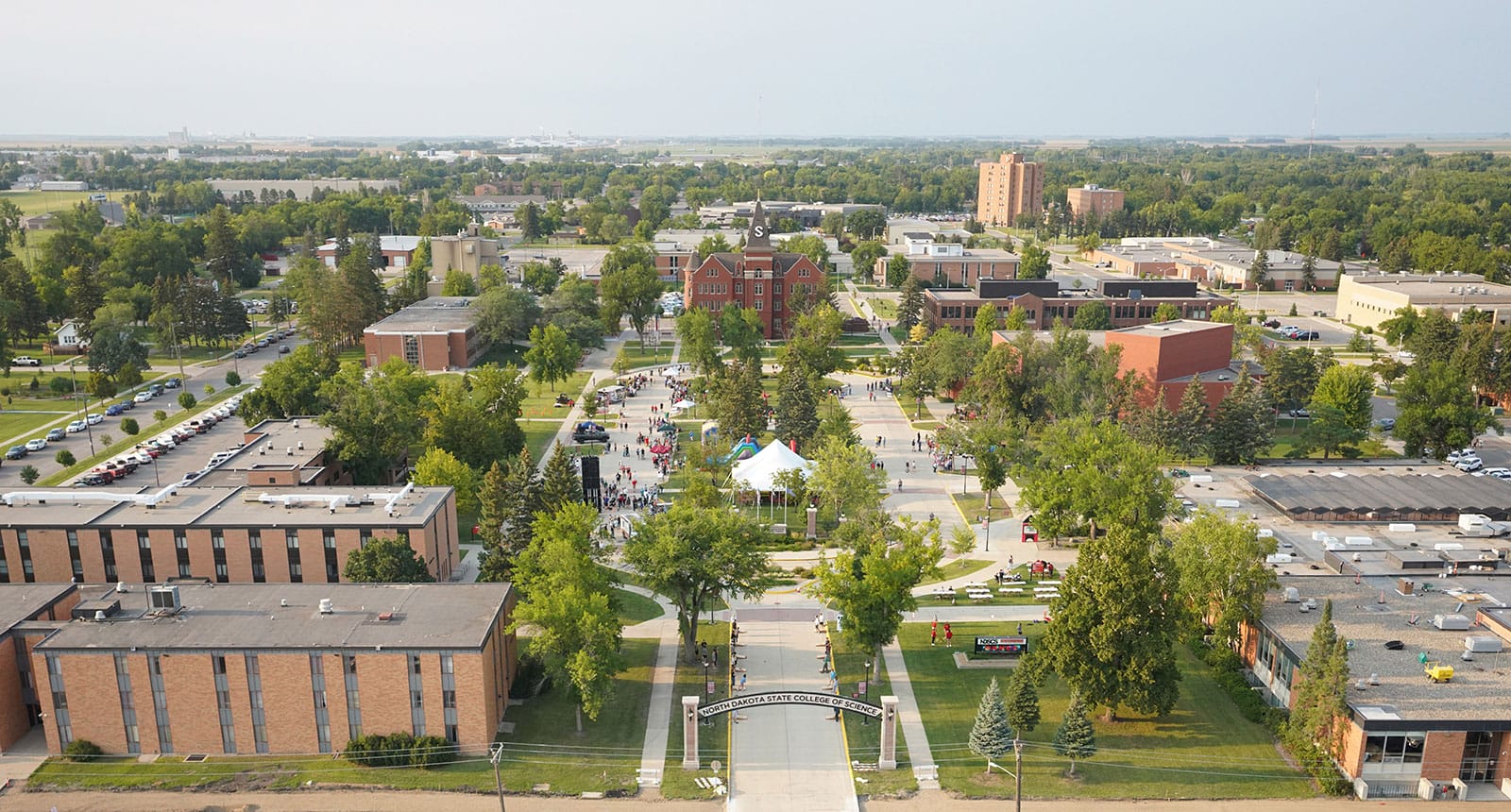 Aerial view of the North Dakota State College of Science campus during an outdoor event, with people gathered around white tents and tree-lined walkways surrounded by academic buildings.