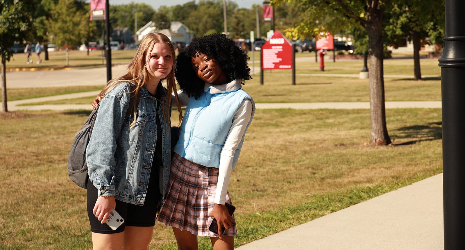 Two students smiling outdoors on campus; one wears a denim jacket and black shorts, the other wears a blue puffer vest and plaid skirt. Trees and signage are visible in the background.
