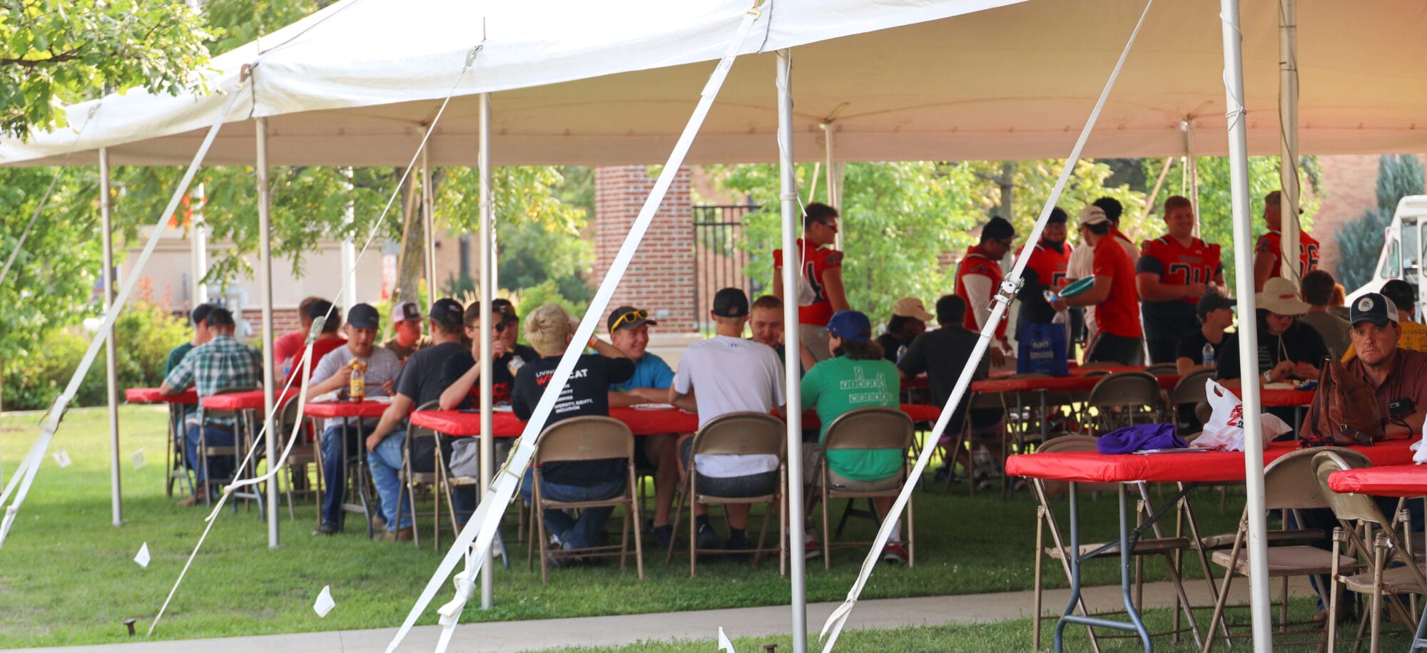 Group of students and athletes gathered under a large outdoor tent with red tablecloths, sitting at folding tables and enjoying a meal or event on a sunny day.