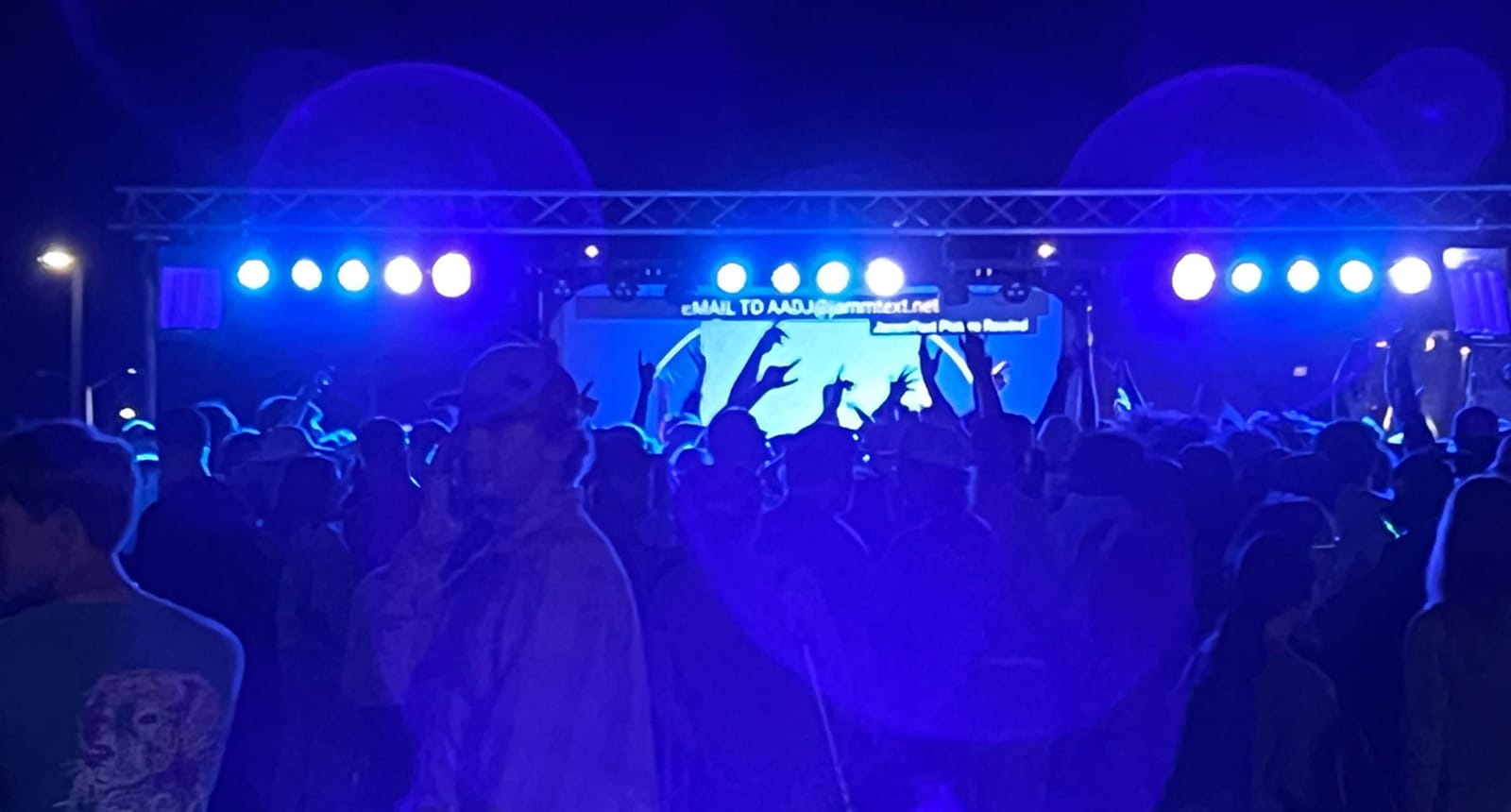 Students gather at an outdoor nighttime concert or dance event, silhouetted by vibrant blue and purple stage lighting.