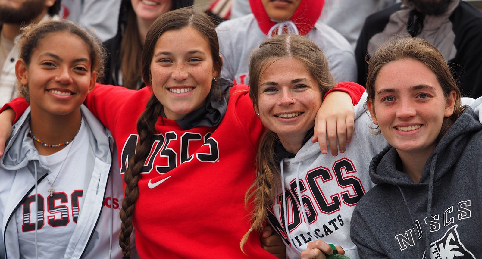Four smiling students wearing NDSCS gear sit closely together, arms around each other, posing for a group photo at an outdoor event.