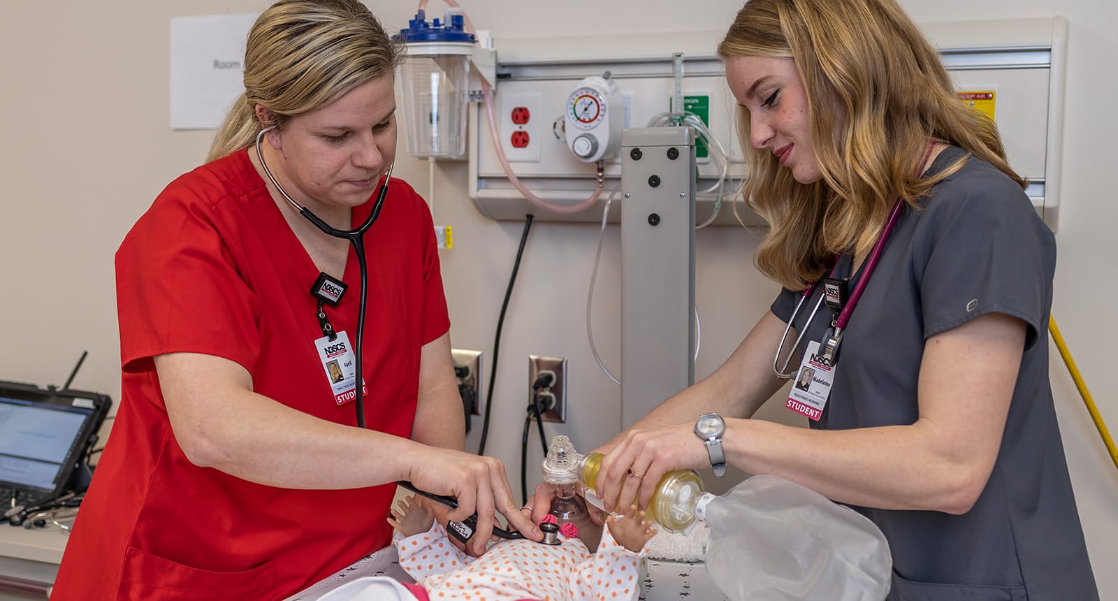 Two nursing students perform a simulated emergency procedure on an infant mannequin using a stethoscope and an ambu bag in a medical training room.