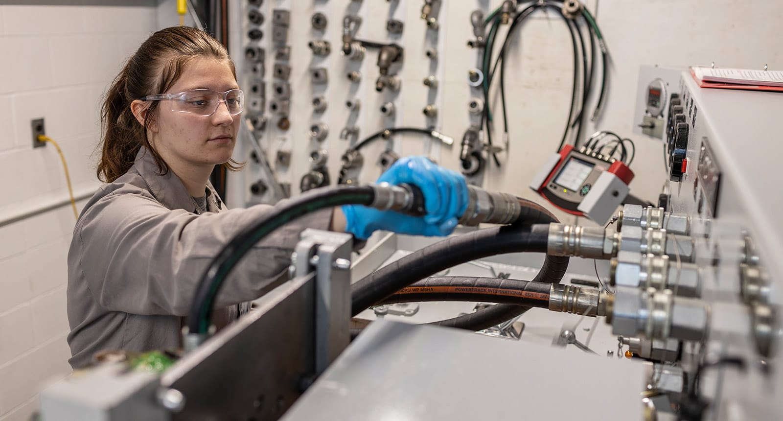 Student wearing safety goggles and gloves connects hydraulic hoses to a testing machine in a technical lab with various fittings and tools on the wall behind her.