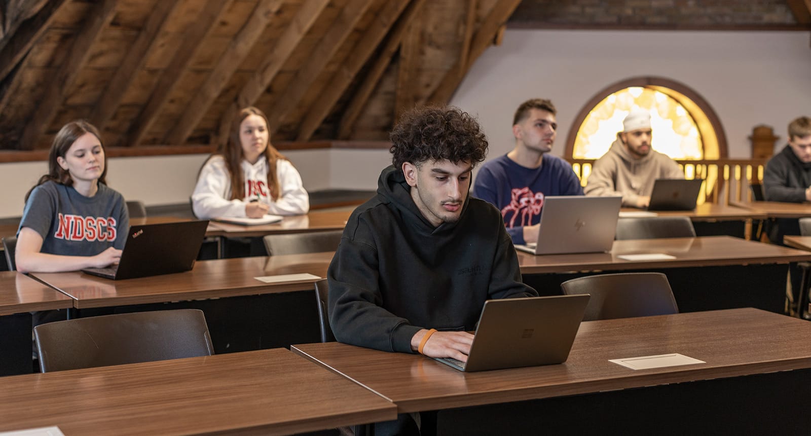 Students sitting at desks in a classroom with laptops, focusing on their work; the classroom has wood-paneled ceilings and a round stained-glass window in the back.