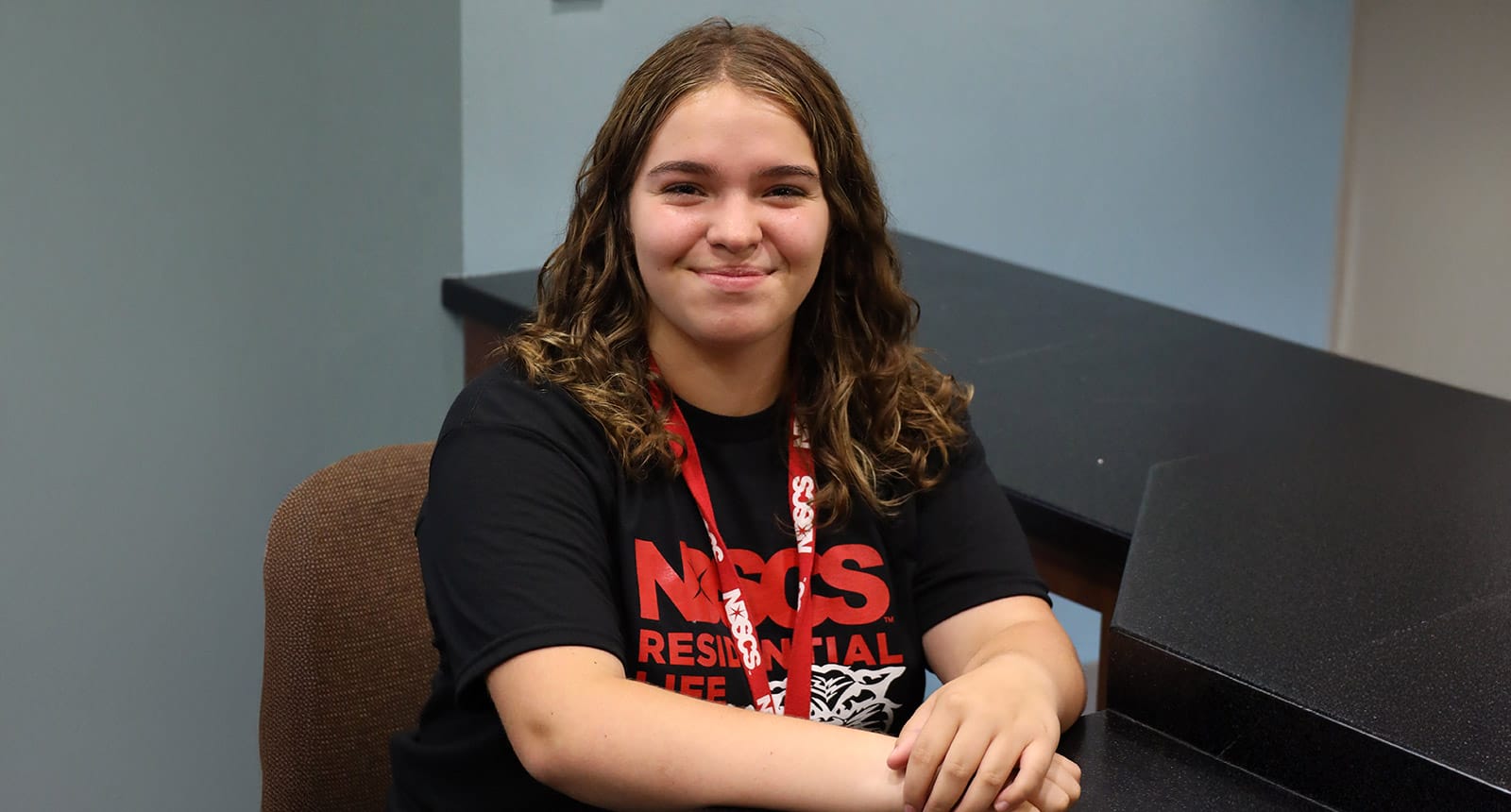 Student smiling while seated at a desk wearing an NDSCS Residential Life t-shirt and lanyard.