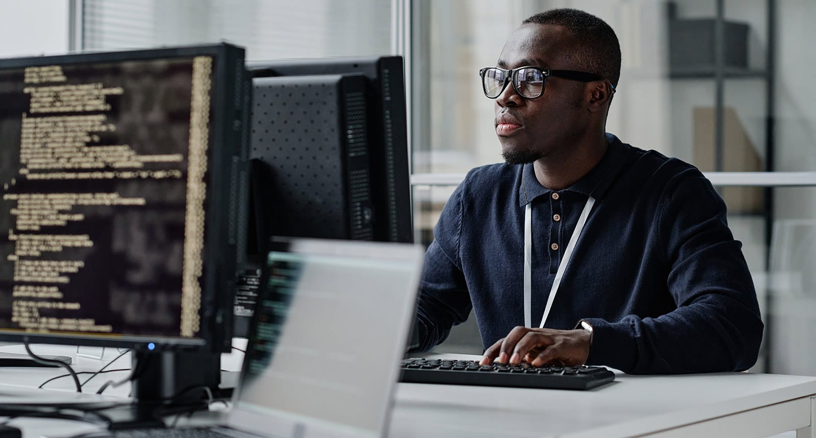 Man wearing glasses and a lanyard focused on a computer screen with code, sitting at a desk in a modern office environment.
