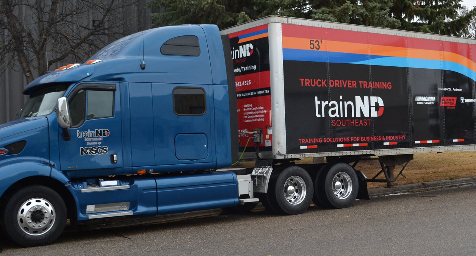 Blue semi-truck and black trailer labeled “trainND Southeast” and “Truck Driver Training,” parked near trees with promotional graphics for training programs and business partnerships.