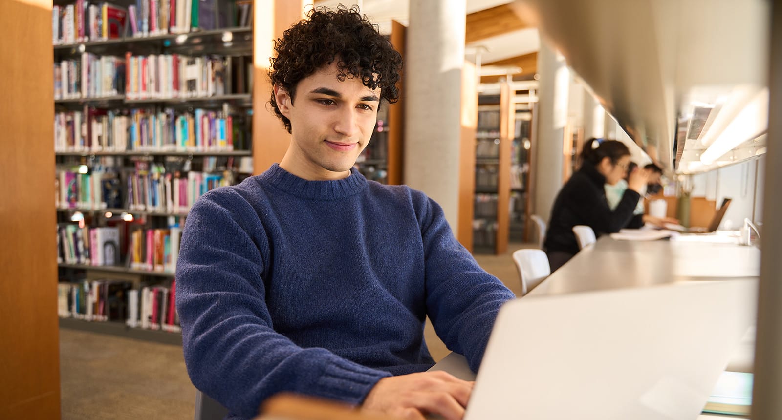 Man at a library looking at his laptop, with bookshelves in the background.