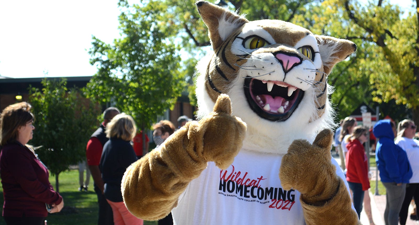 NDSCS wildcat mascot wearing a “Wildcat Homecoming 2021” t-shirt, smiling and giving two thumbs up at an outdoor campus event with people gathered in the background.