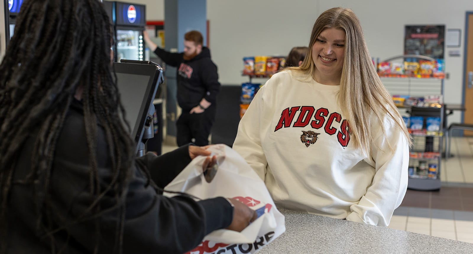Smiling student in a white NDSCS sweatshirt standing at a campus store counter as another person hands her a shopping bag.