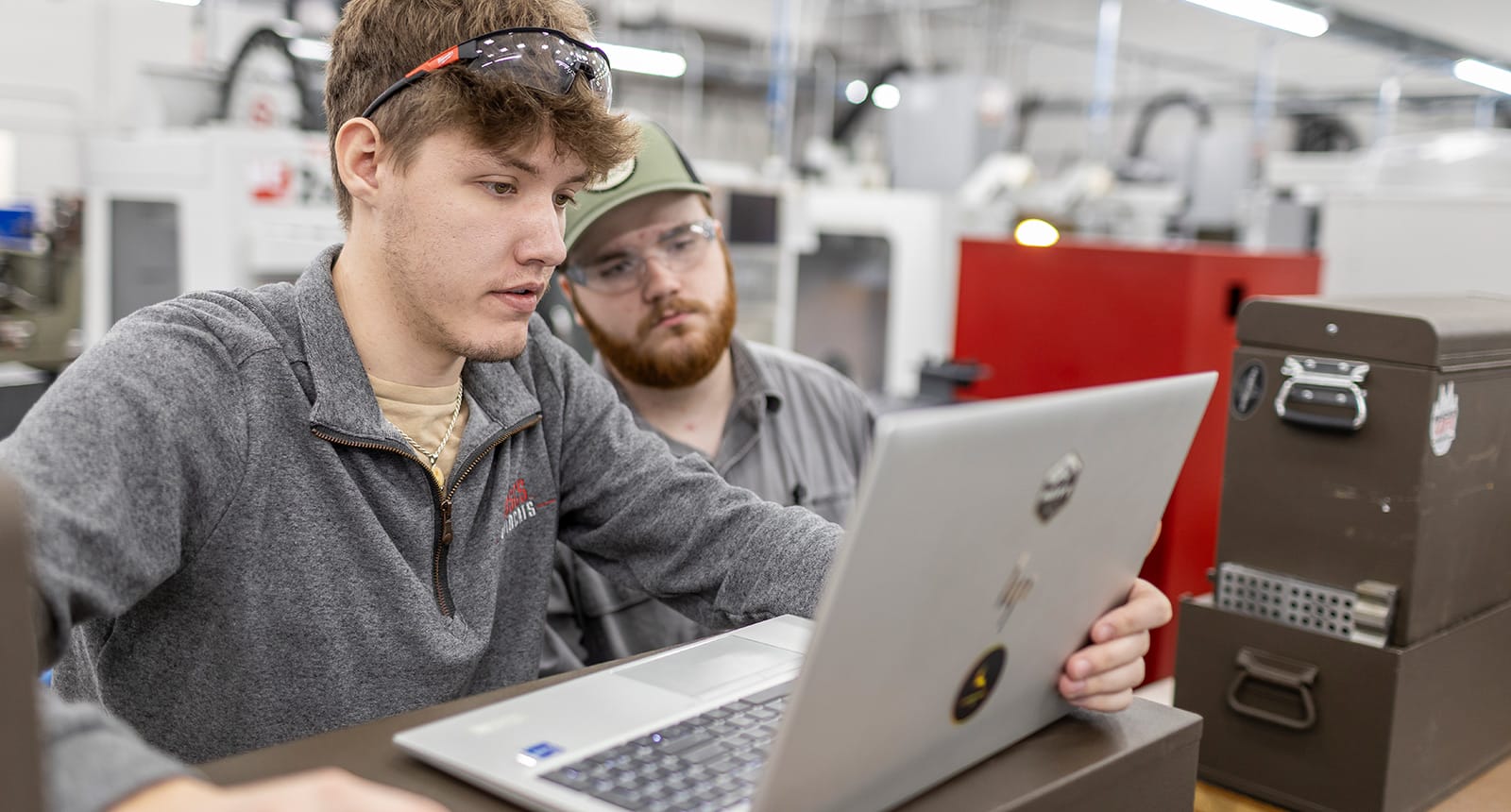 Two male students in a workshop setting looking intently at a laptop; one is pointing at the screen while the other observes, surrounded by industrial equipment and toolboxes.