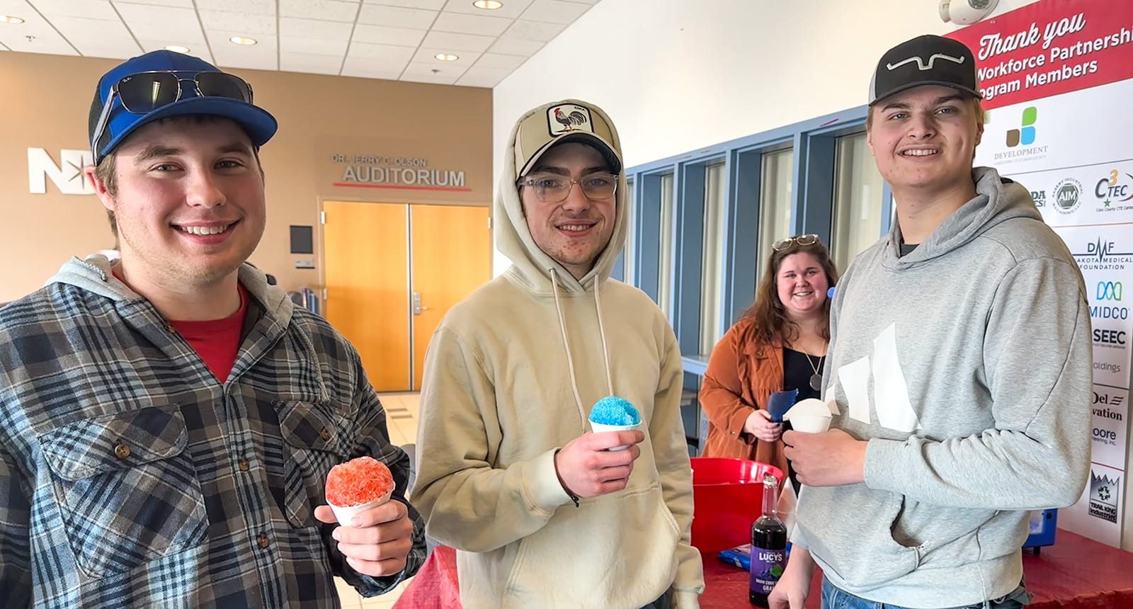 Three smiling students hold colorful snow cones in a campus lobby, standing in front of a Workforce Partnership banner and the entrance to an auditorium.
