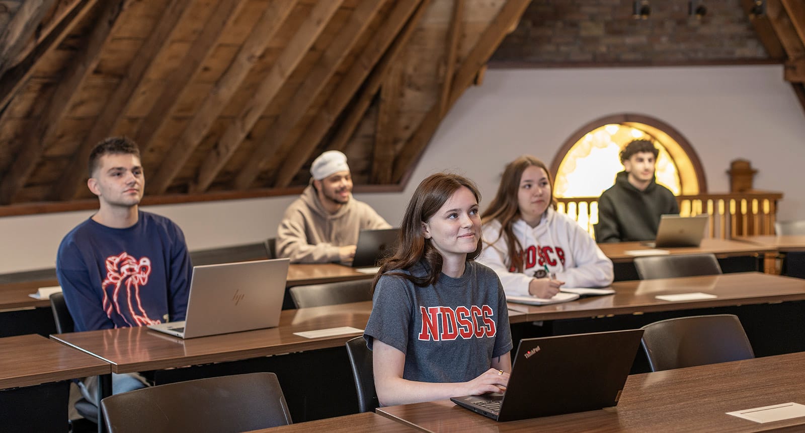 Five students sitting at desks with laptops in a rustic classroom with wooden beams and a stained glass window, attentively listening.