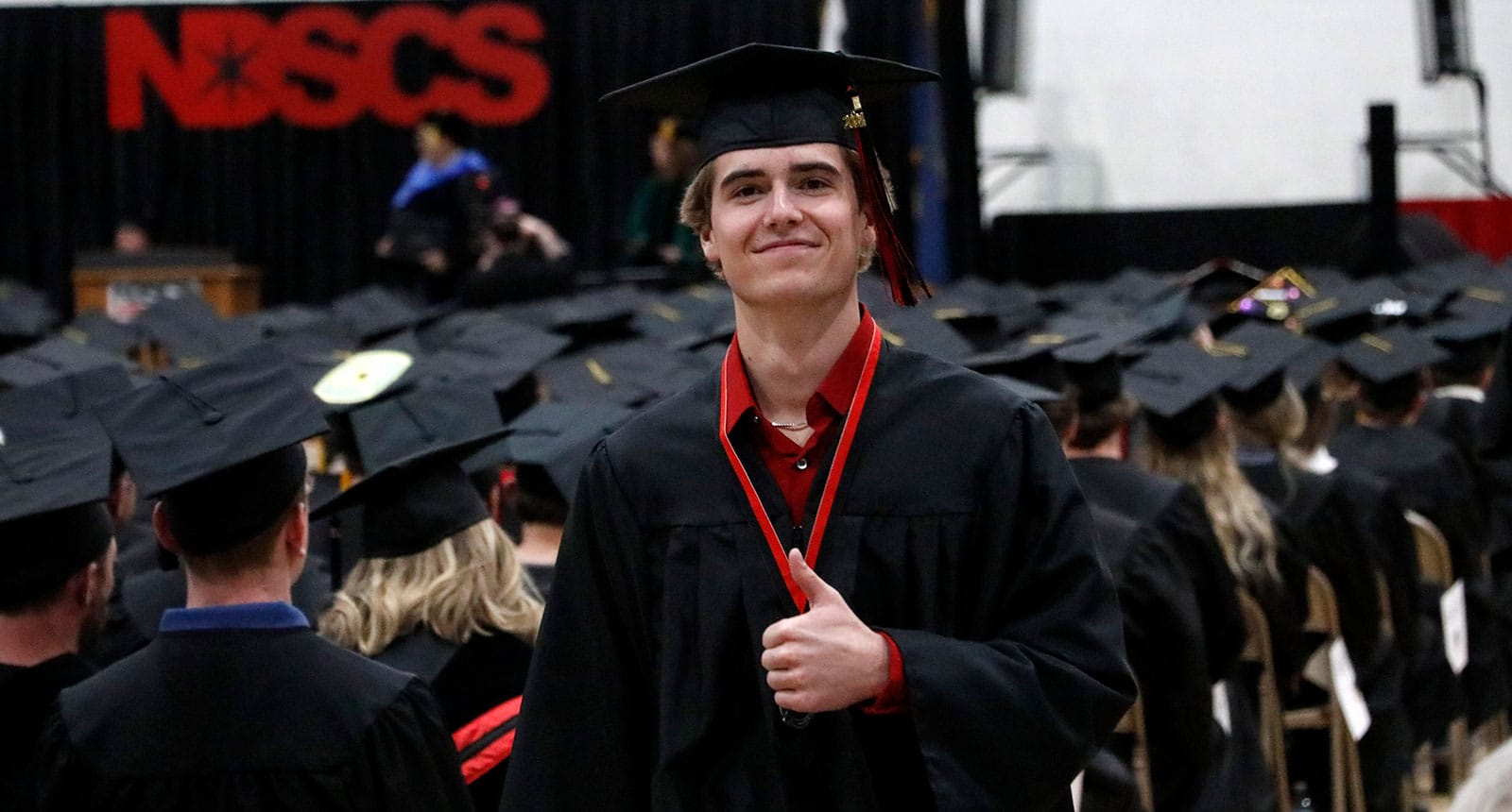 Smiling graduate in a black cap and gown gives a thumbs-up during a commencement ceremony at NDSCS, standing among a large group of seated graduates.
