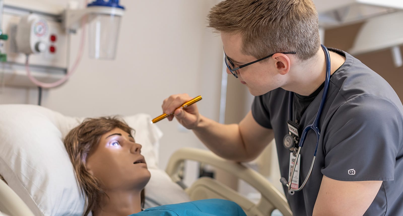 Nursing student in scrubs using a penlight to check the pupil response of a medical training mannequin lying in a hospital bed.