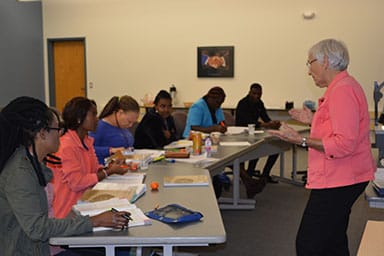 Older woman in pink shirt teaching a diverse group of adult students seated at classroom tables with open books and study materials.