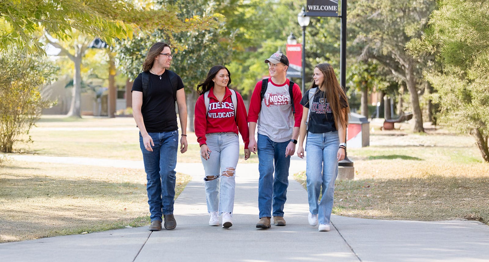 Four students walking together on a tree-lined campus path, smiling and chatting, wearing NDSCS Wildcats shirts and carrying backpacks.