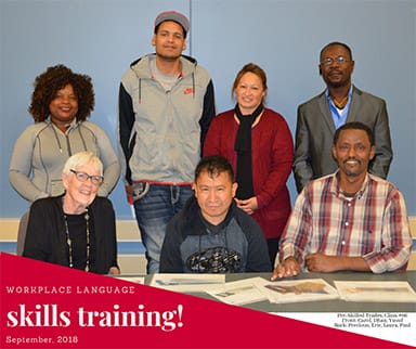 Group of eight adults posing in a classroom setting for a photo labeled “Workplace Language Skills Training – September 2018.” Some are seated at a table with papers while others stand behind them, all smiling.