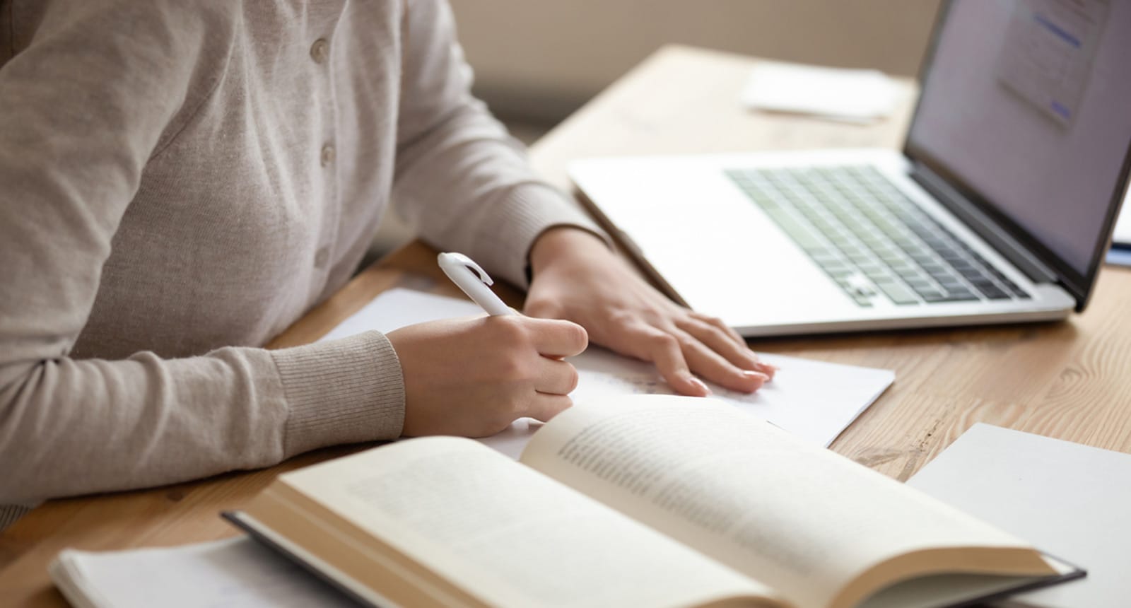 Person writing in a notebook with an open book and laptop on a wooden desk, suggesting a study or online learning environment.