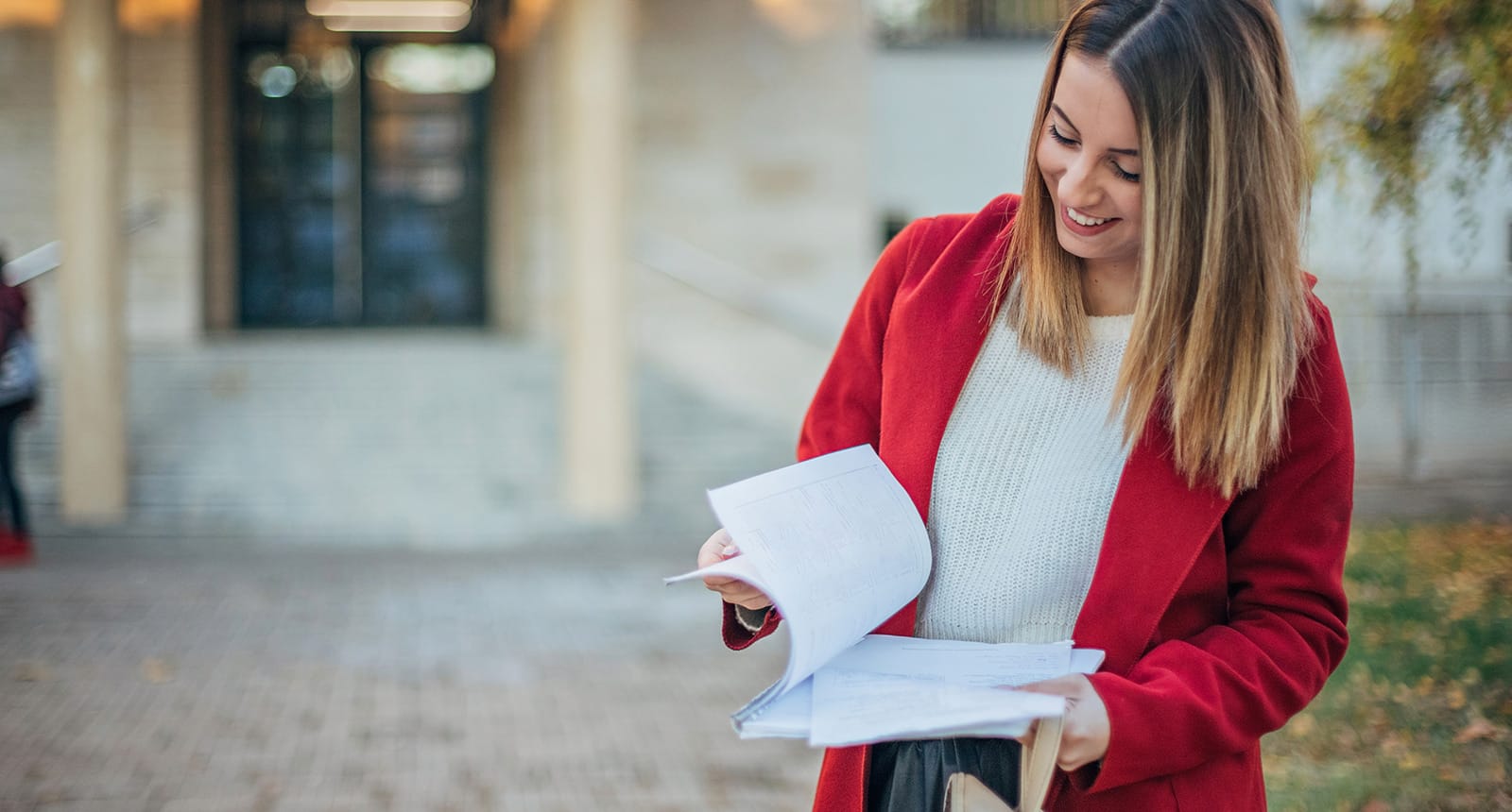 Smiling young woman with straight hair wearing a red coat and white sweater, flipping through papers outdoors in front of a building entrance.