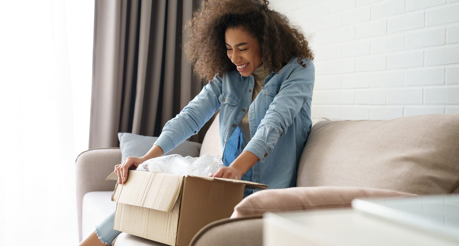 Smiling woman with curly hair sitting on a beige couch and excitedly opening a cardboard box filled with bubble wrap in a bright living room.