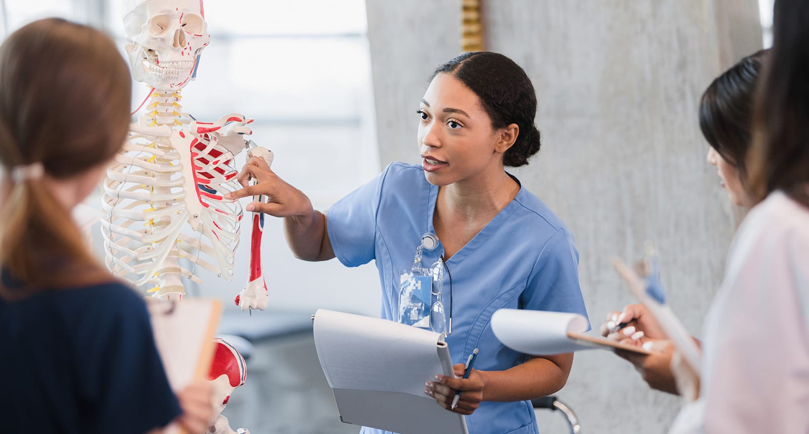 Medical student in blue scrubs explaining parts of a human skeleton model to classmates holding clipboards.