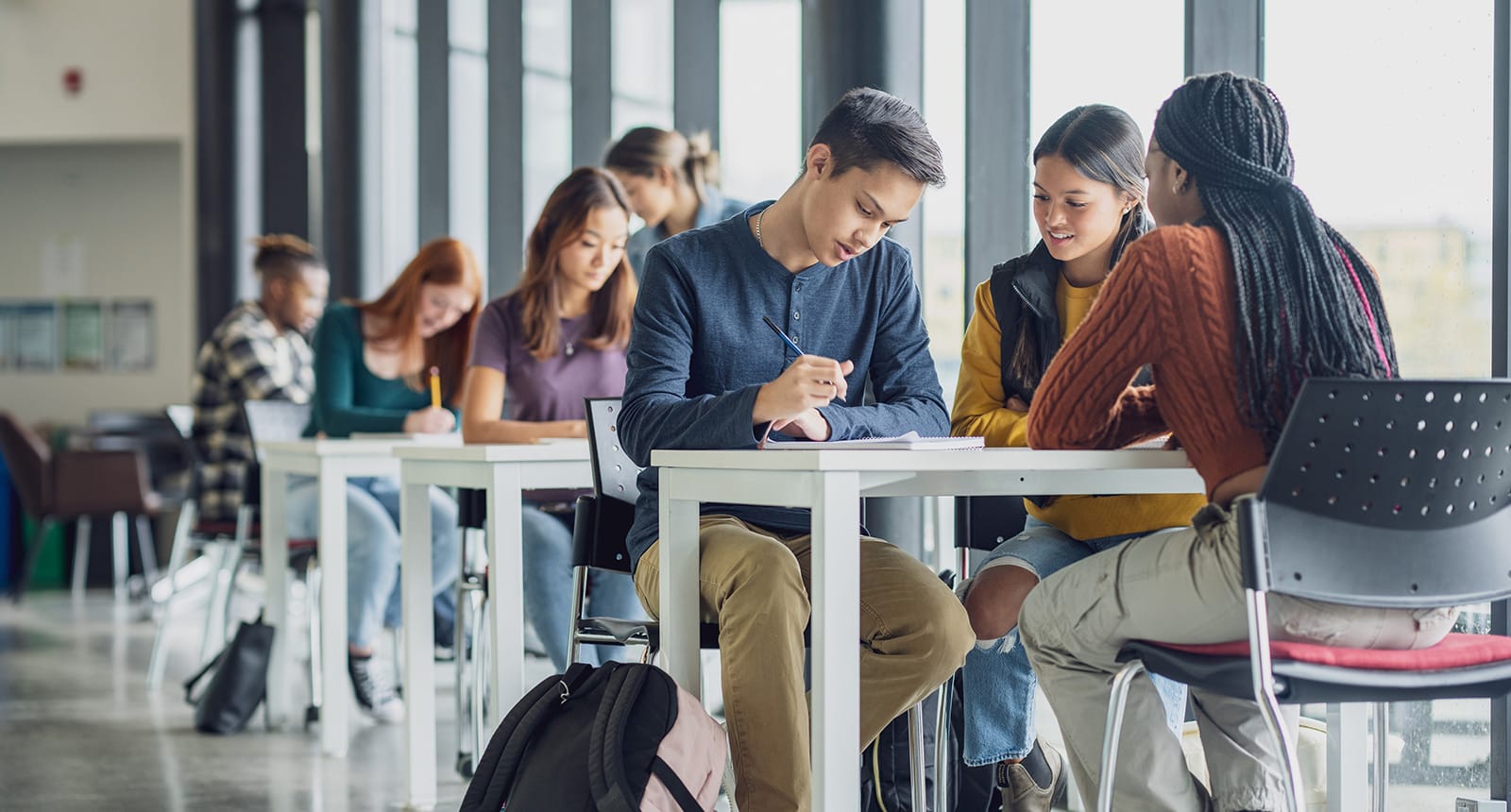Group of high school or college students working together at tables in a bright, modern study area with large windows, notebooks and backpacks visible.