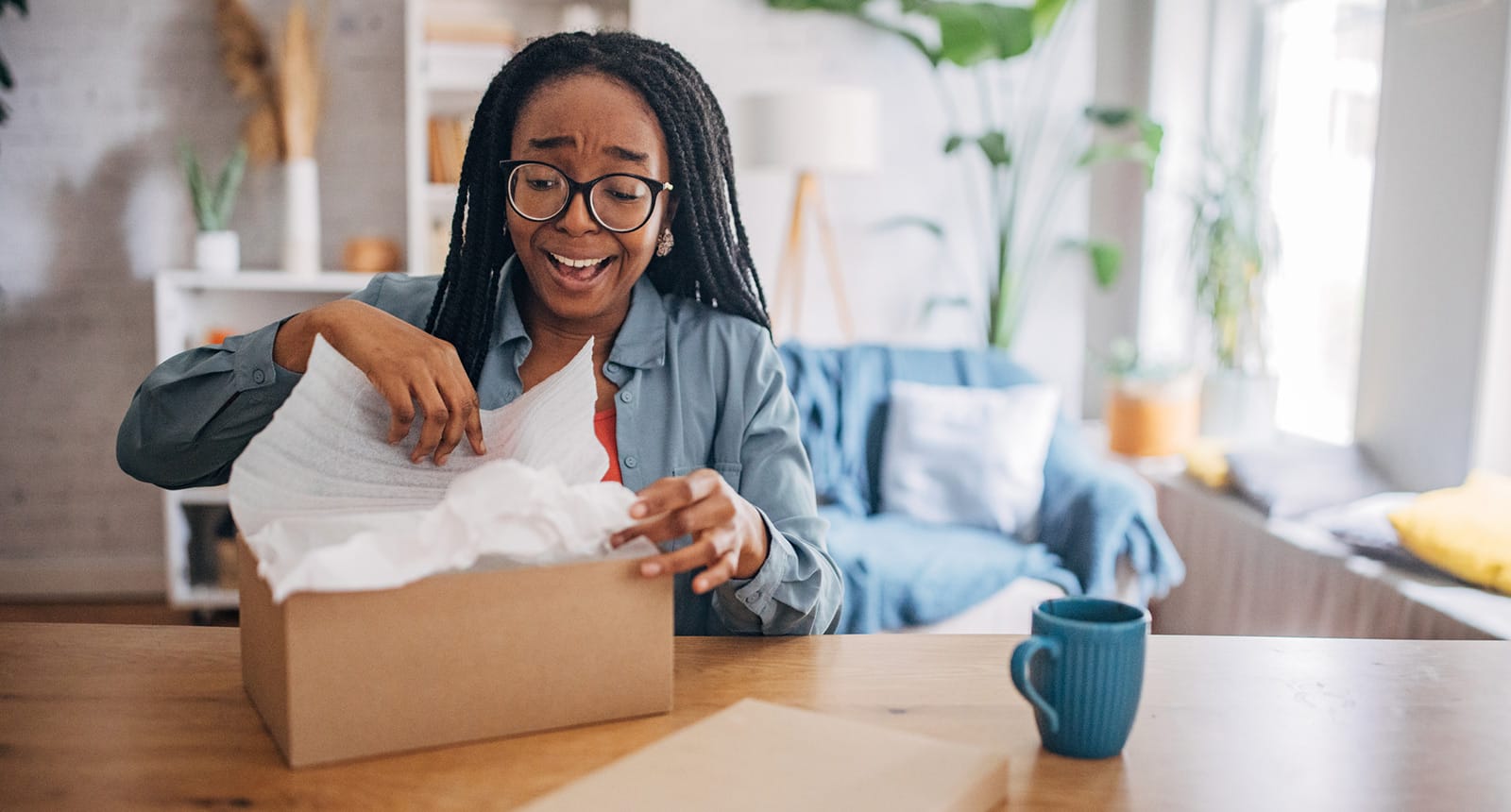 Smiling woman with long braids and glasses opening a cardboard box with excitement while sitting at a wooden table in a cozy, brightly lit living room.