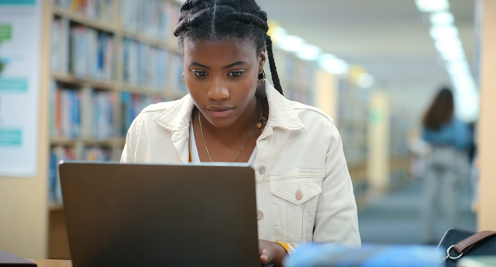 Young woman with braids wearing a light-colored jacket focused on a laptop in a brightly lit library aisle.