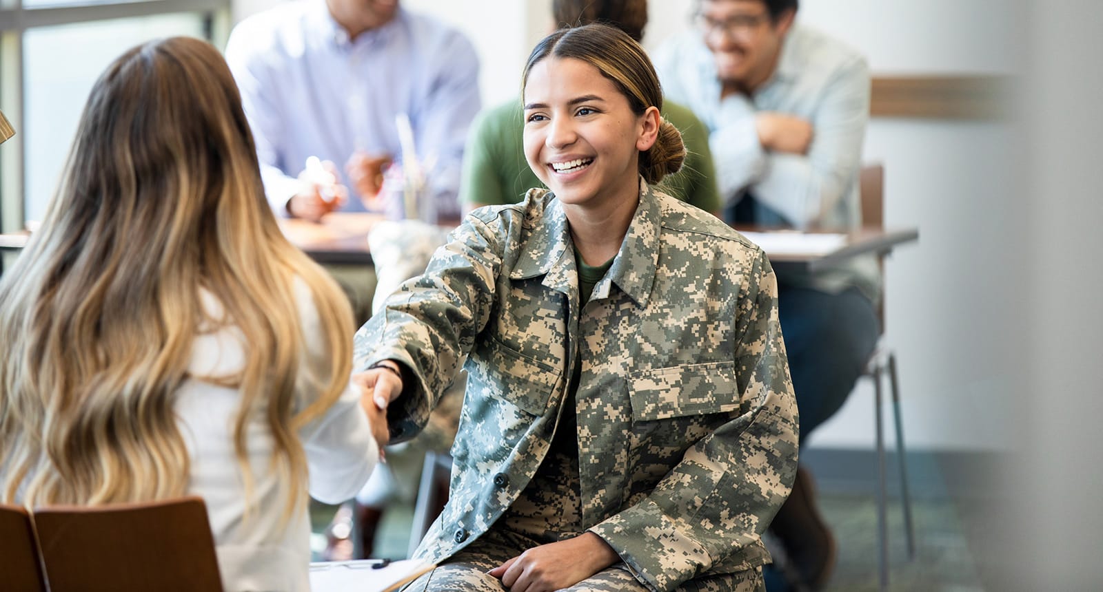 Smiling young woman in military camouflage uniform shaking hands with another woman across a table in a classroom or meeting setting, with others talking in the background.