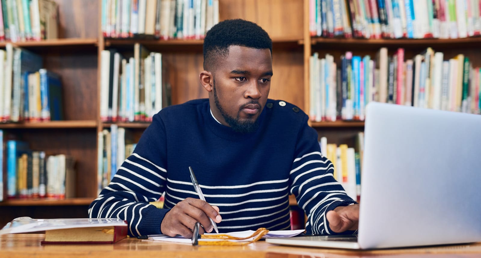 Focused student in a navy striped sweater working on a laptop and taking notes in a library with bookshelves in the background.