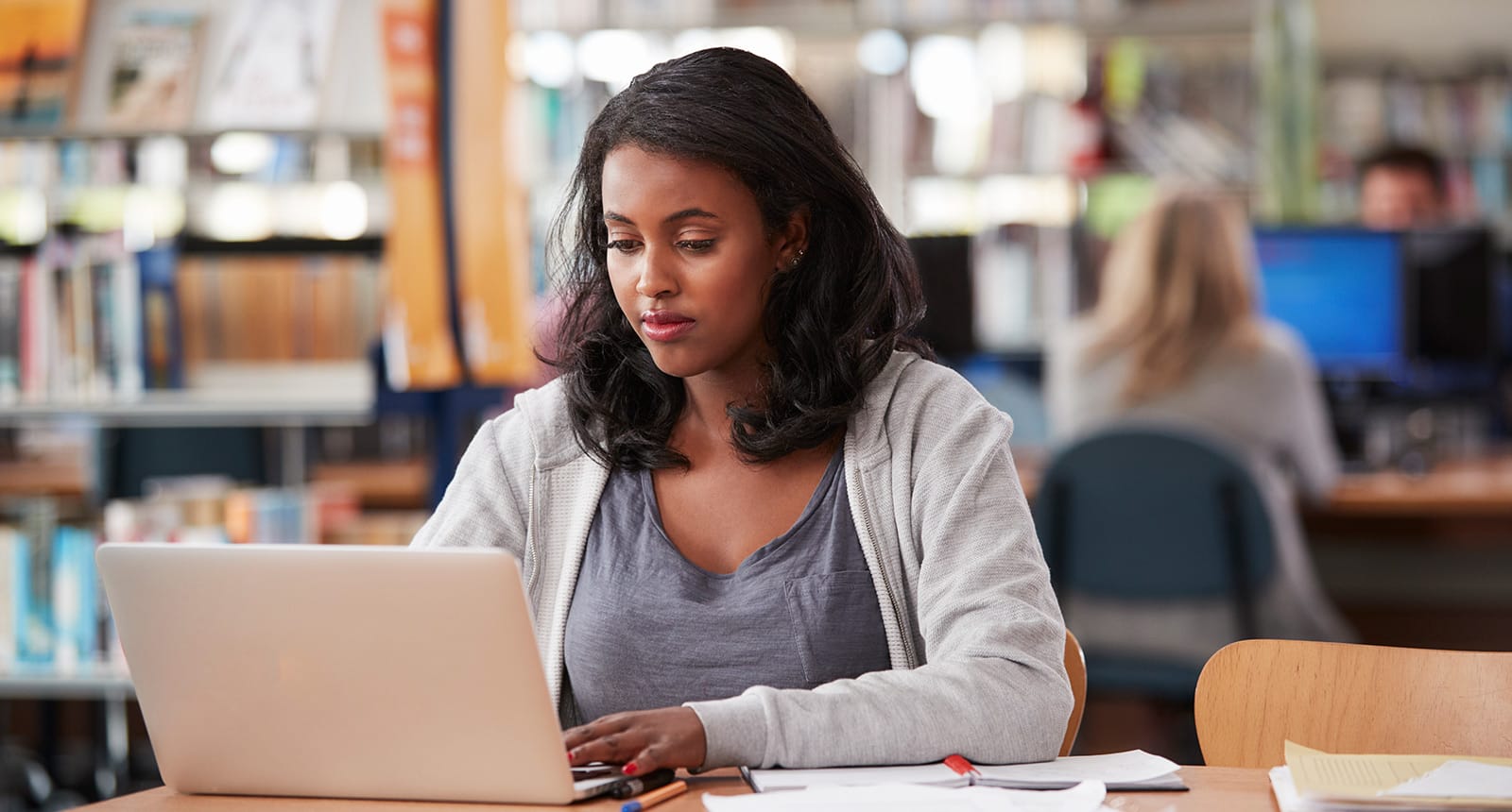 Focused woman sitting at a desk in a library, working on a laptop with notebooks and pens spread out in front of her.
