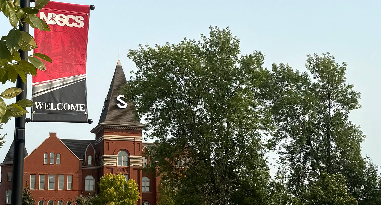 Brick academic building with a pointed tower marked “S” partially obscured by trees, next to a red and black NDSCS “Welcome” banner on a lamppost.