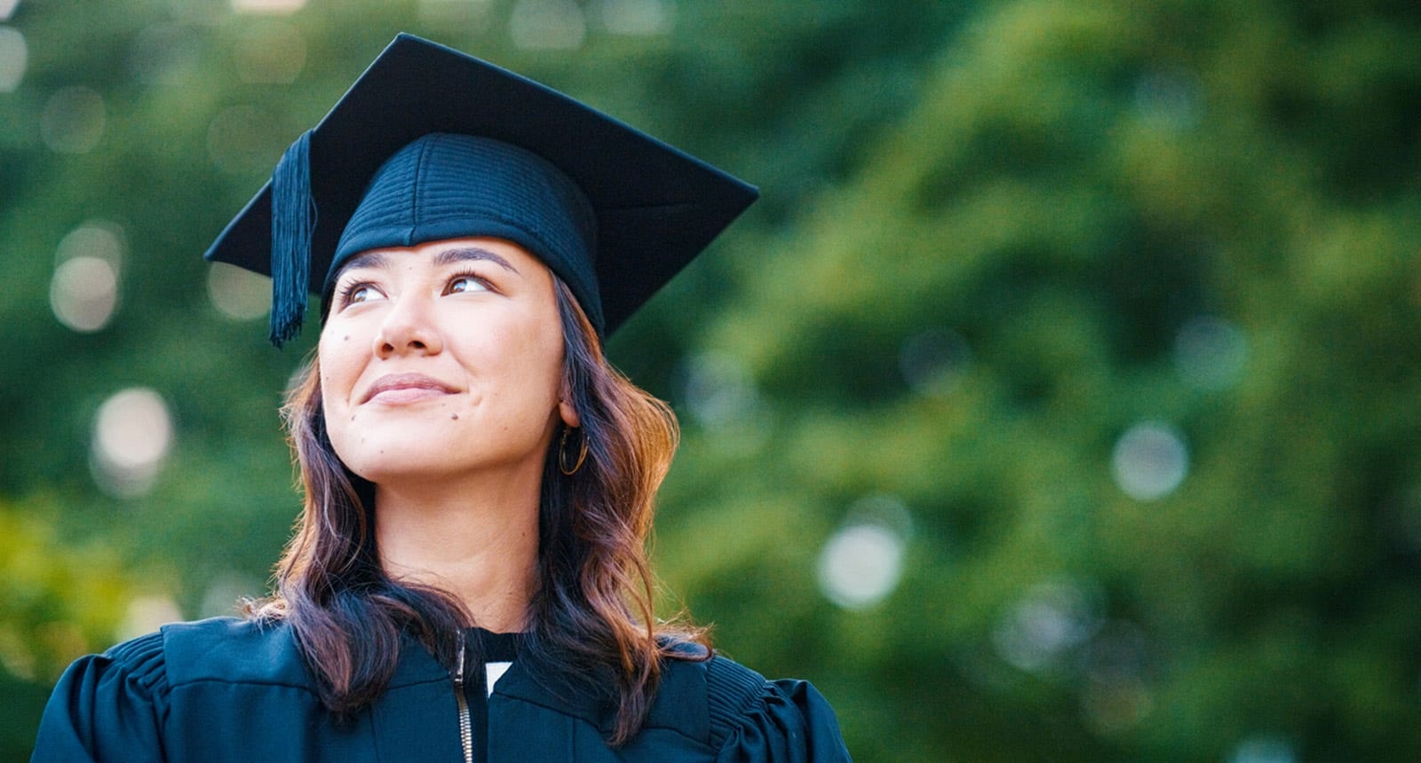 Smiling young woman in a black graduation cap and gown looking upward with blurred green trees in the background.