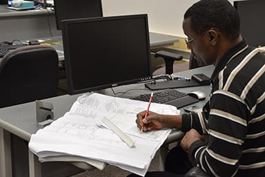Man seated at a desk in a computer lab, reviewing and marking up large engineering or architectural plans with a red pencil and ruler.