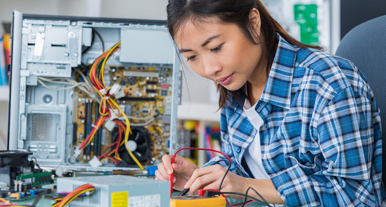 Student in a blue plaid shirt using a multimeter to test computer hardware components beside an open desktop tower with visible wiring and circuit boards.