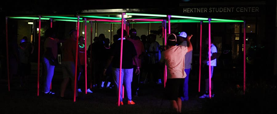 Students participate in a nighttime glow game under blacklight, surrounded by neon pink and green poles, outside the Hektner Student Center.