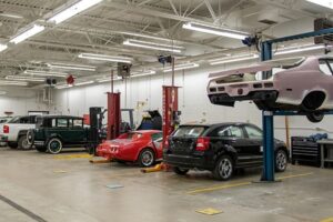 Automotive workshop with multiple cars in various stages of repair, including one car lifted on a hydraulic lift and others parked at workstations.