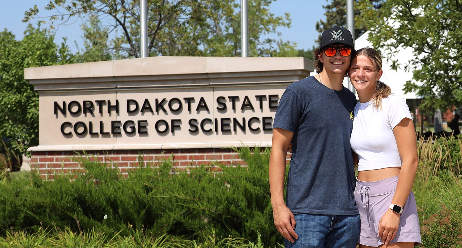 Two smiling students standing in front of a North Dakota State College of Science sign on a sunny day.