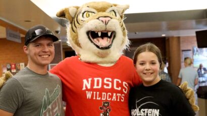 Two students pose with the NDSCS Wildcats mascot, who is wearing a red school shirt, inside a campus building.