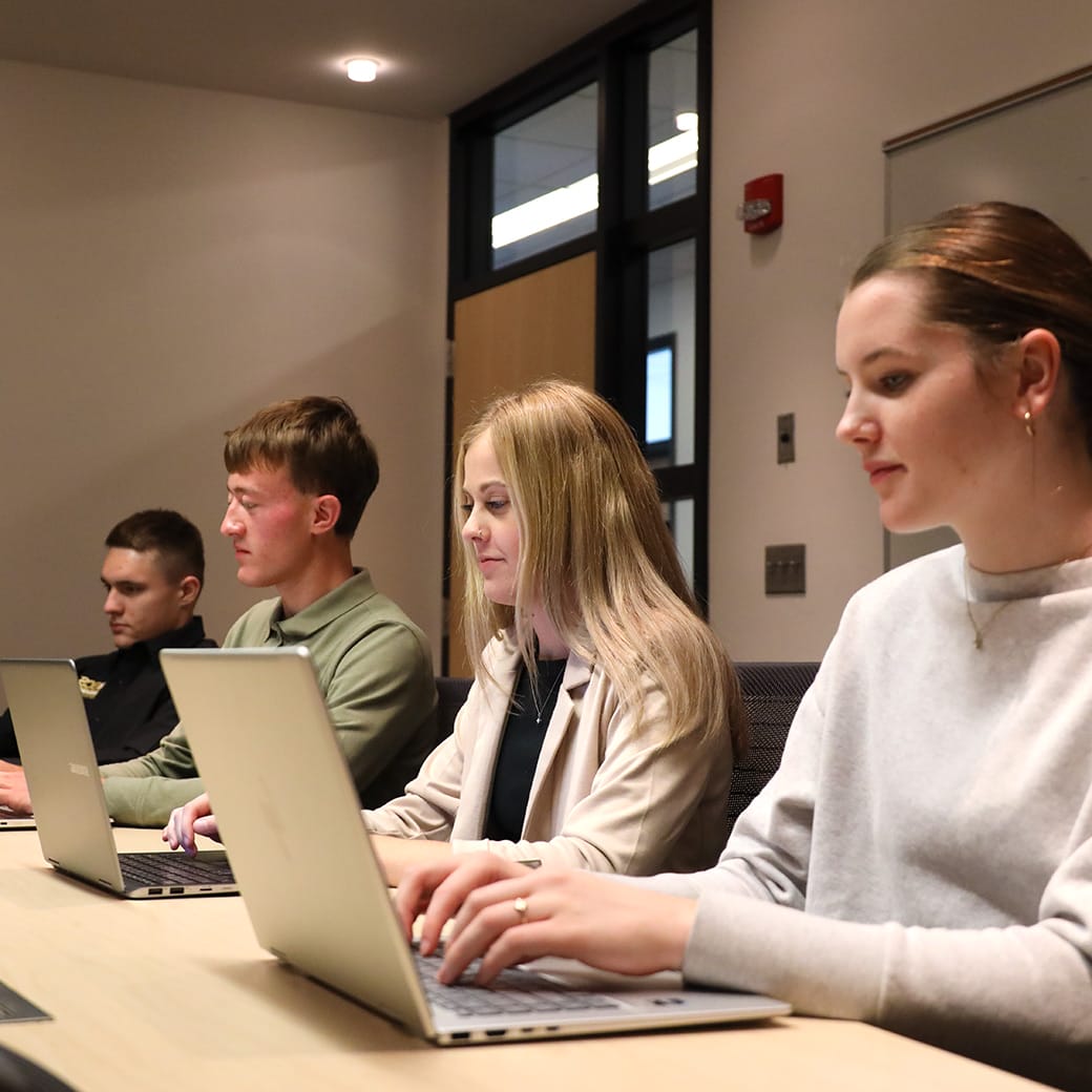Four students working on laptops in a classroom setting, seated side-by-side at a long table, focused on their screens.