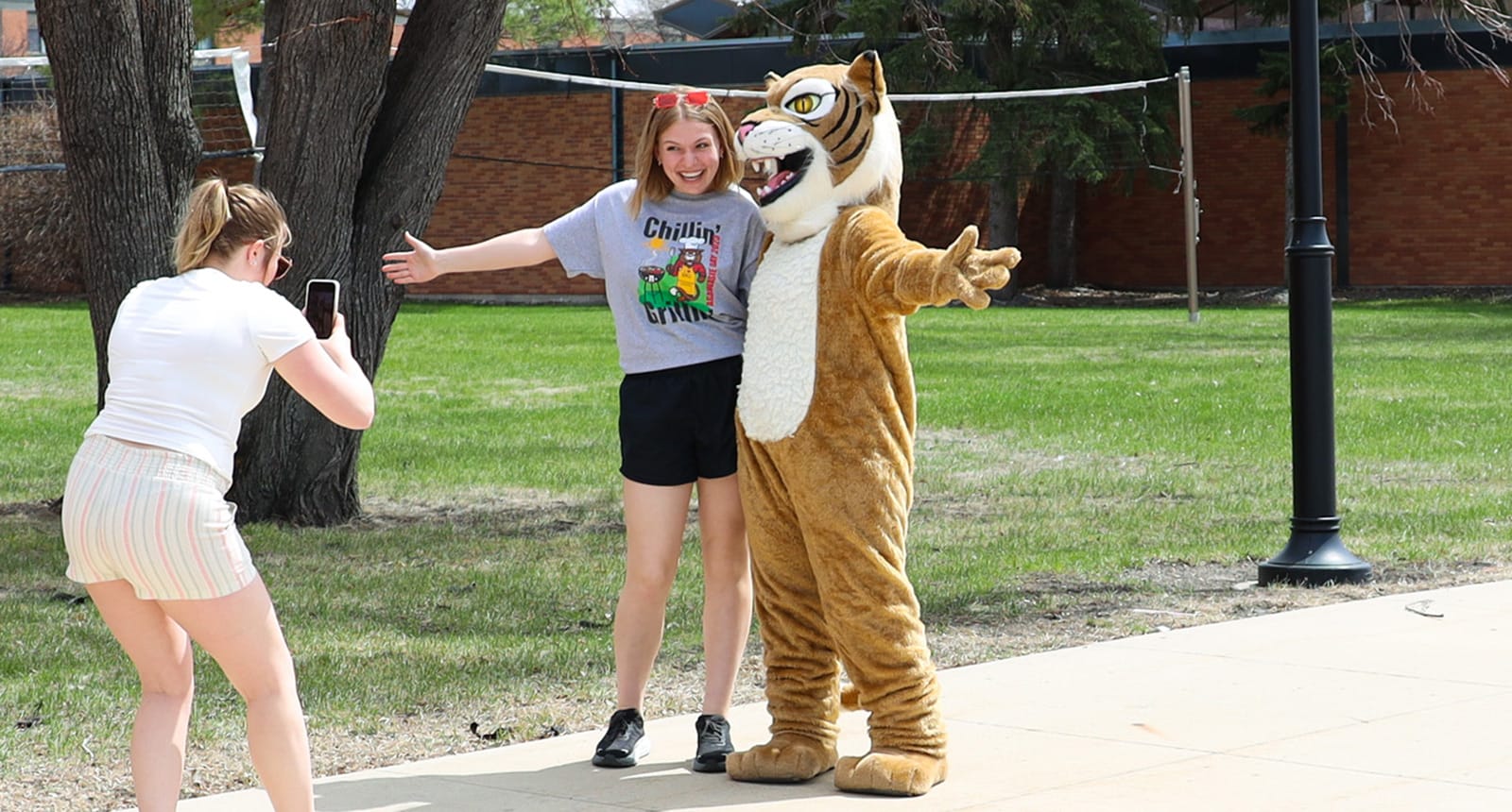 A student poses outdoors with the NDSCS Wildcat mascot, arms wide and smiling, while another student takes their photo with a smartphone.