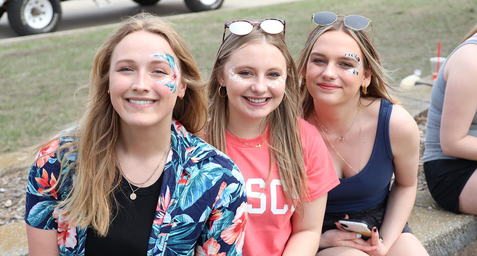 Three young women sitting outdoors, smiling, with colorful face paint and casual summer clothing.