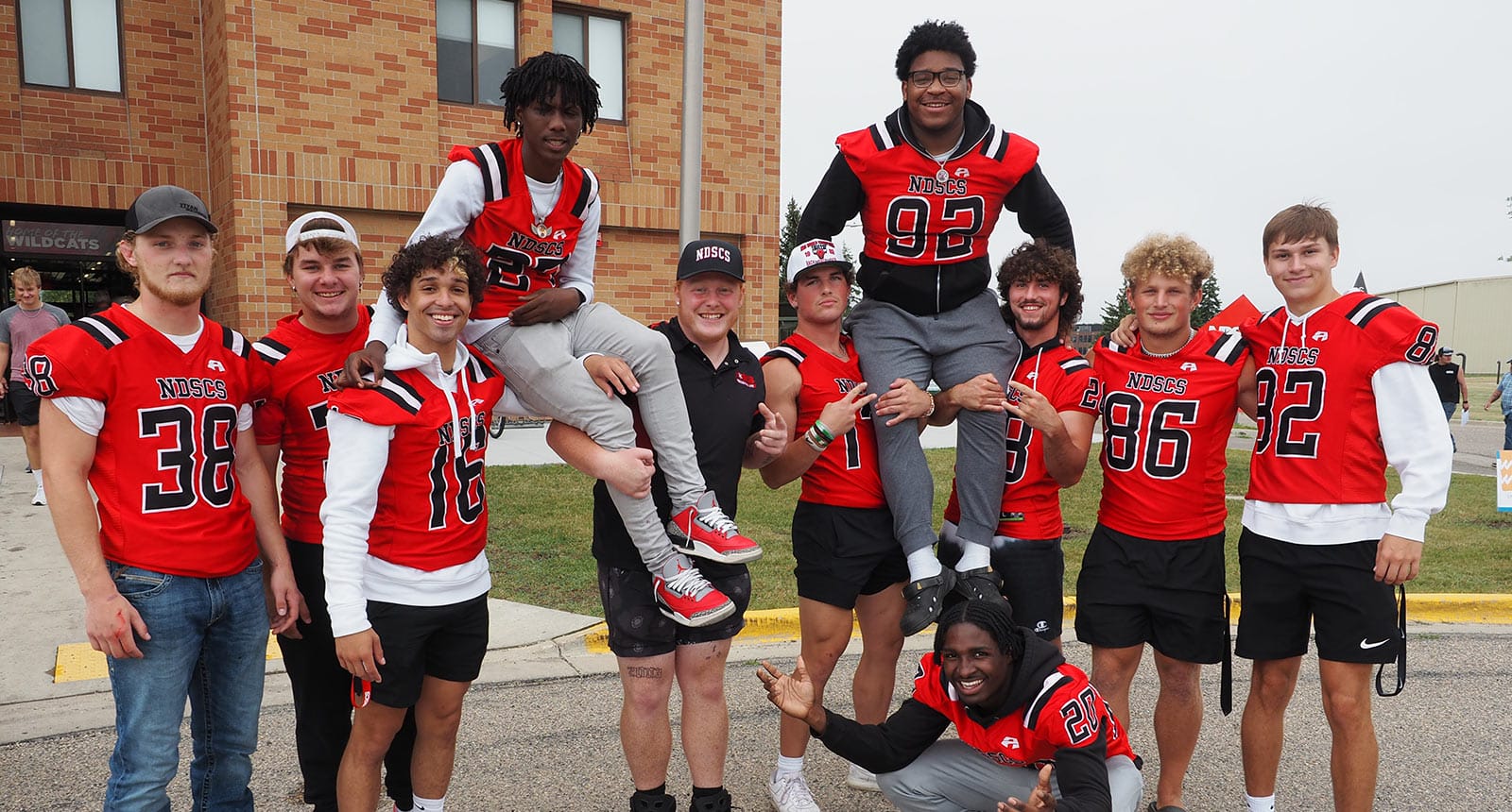 Group of NDSCS football players in red and black jerseys posing together outside a campus building, with two teammates being lifted on shoulders and everyone smiling or making playful gestures.