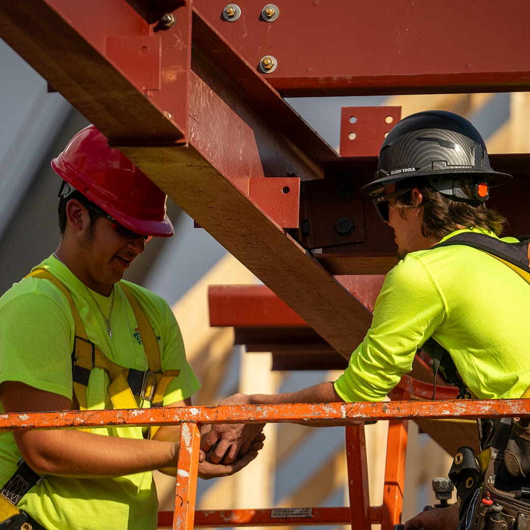 Two construction workers in bright safety gear and hard hats working on a steel beam structure while standing on an orange lift.