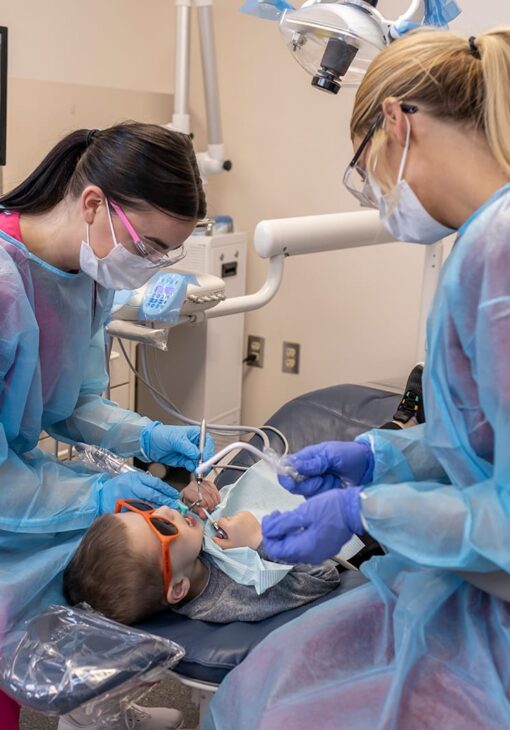 Two dental technicians in masks and protective gowns work on a young child wearing orange sunglasses, who is reclined in a dental chair during a procedure.