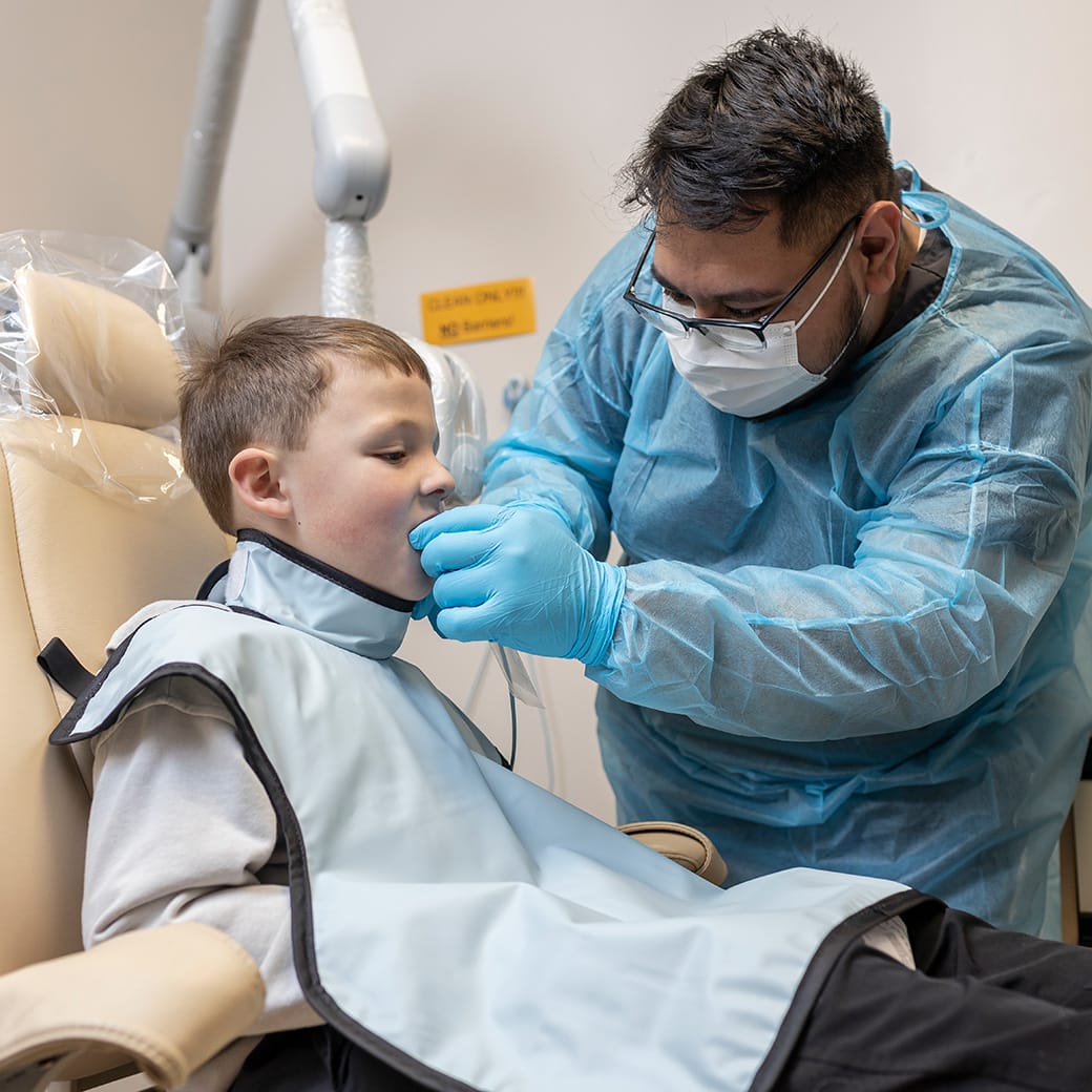 A dental technician in a mask and protective gown assists a young boy seated in a dental chair during an imaging procedure, both focused on the process.