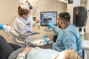 Two dental professionals review a patient’s dental X-ray on a computer monitor while the patient lies reclined in a dental chair.