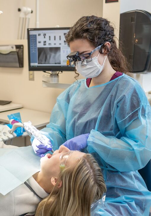 Dental hygienist wearing magnifying loupes and protective gear examines a female patient’s teeth while an X-ray is displayed on the monitor in the background.